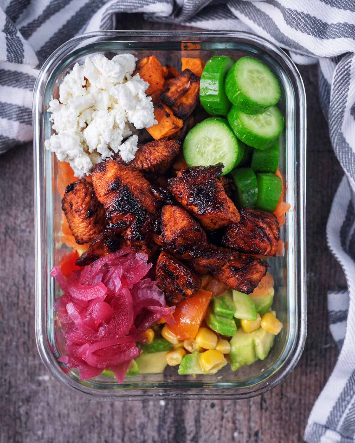 A glass storage container full of cooked chicken, salad items and crumbled feta.