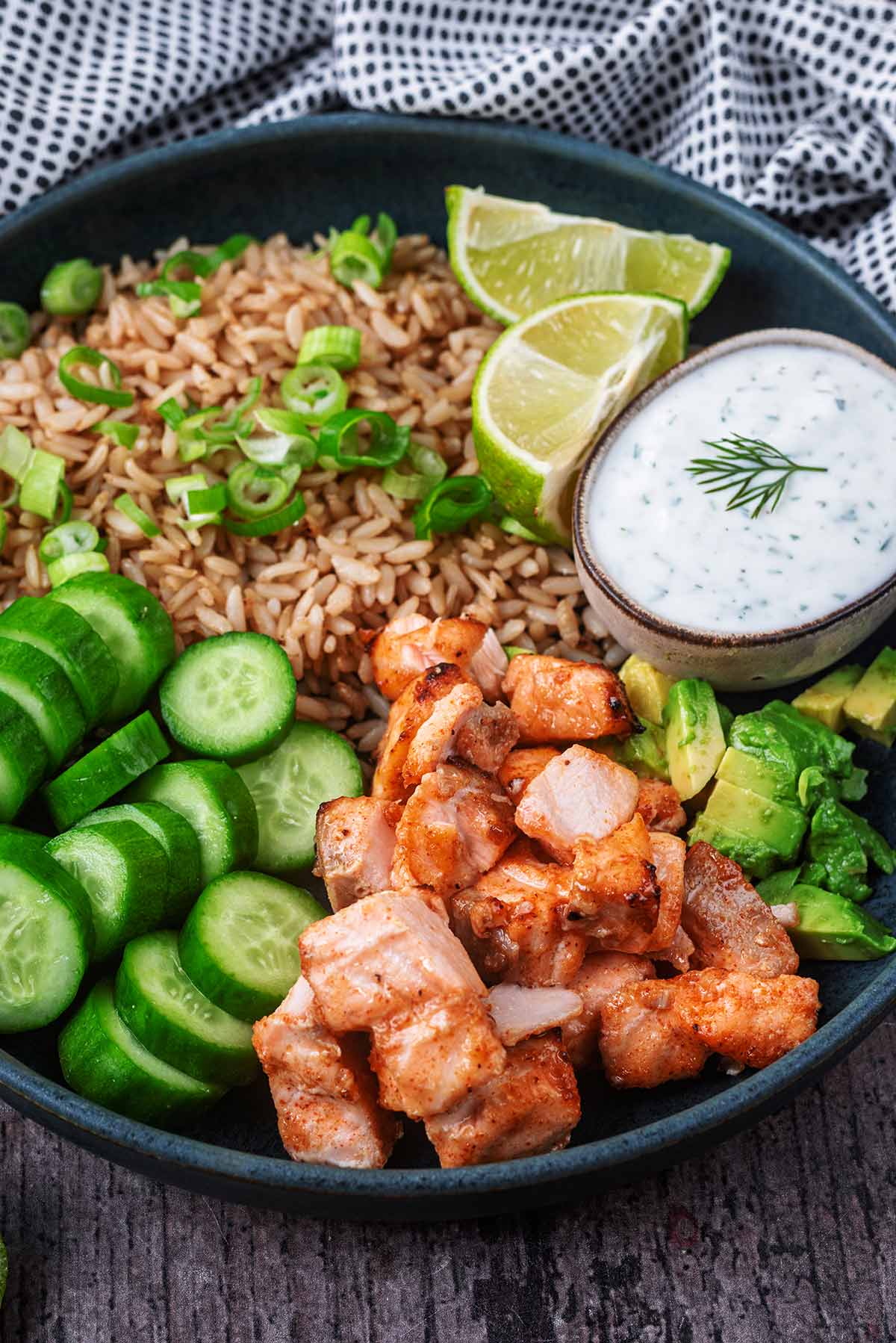 A bowl containing, cooked salmon bites, rice, cucumber slices, diced avocado and a small pot of creamy dip.