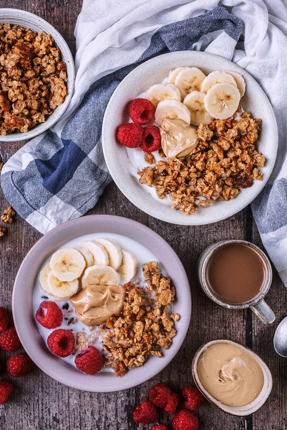 Two bowls of scrambled oats, banana slices and raspberries. One bowl has yogurt, the other has milk.