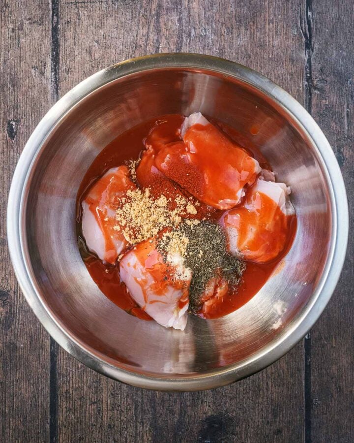 Raw chicken thighs in a mixing bowl with hot sauce and spices.