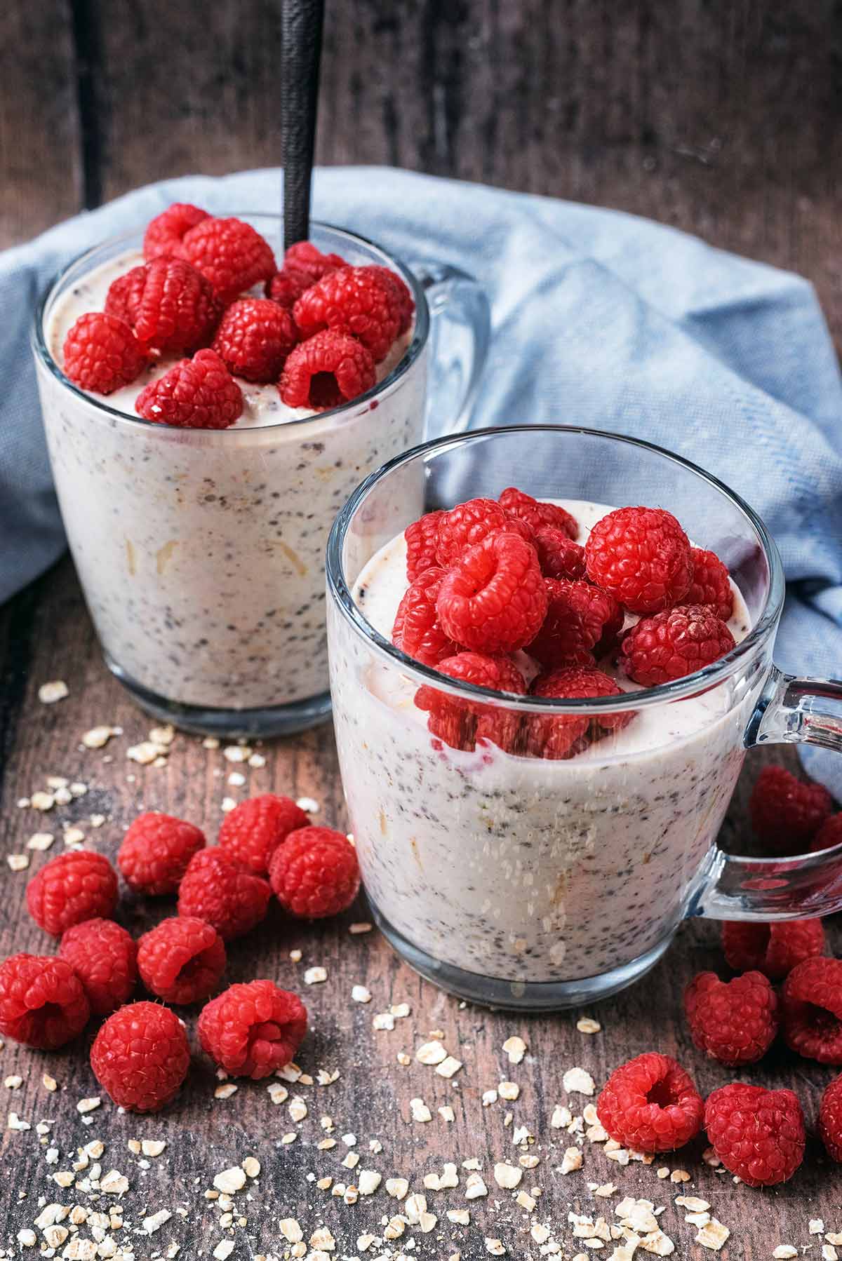 Two serving glasses full of overnight oats, surrounded by raspberries and oats.