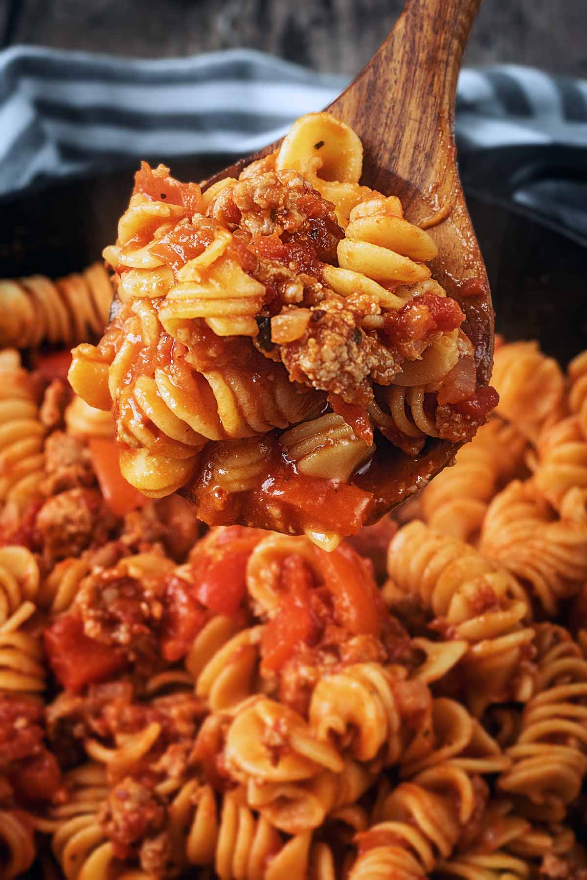 A wooden spoon lifting some sausage pasta out of a pan.