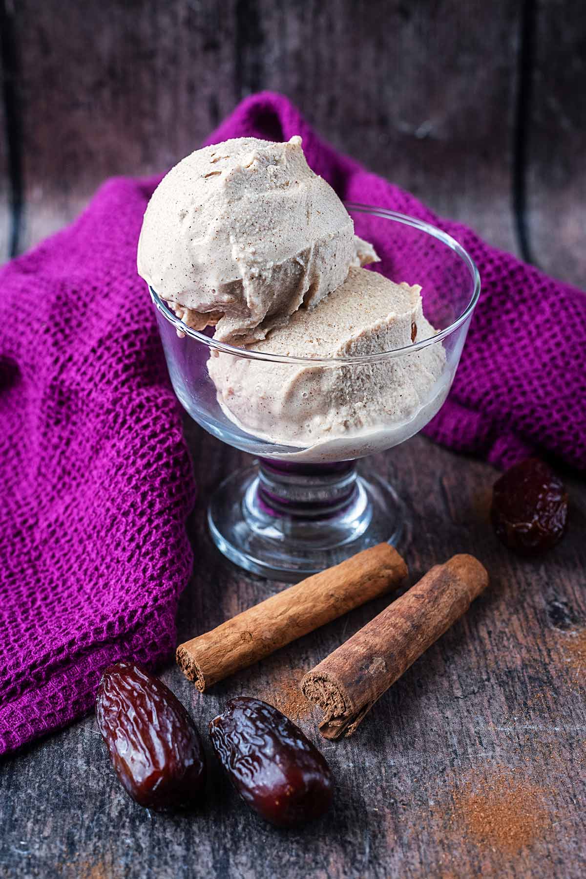 Two balls of ice cream in a serving glass next to some dates and cinnamon sticks.