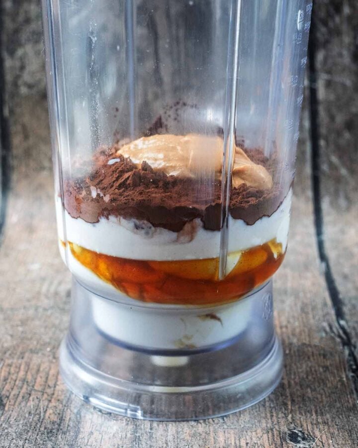 A blender jug containing yogurt, milk, maple syrup, cocoa and peanut butter.