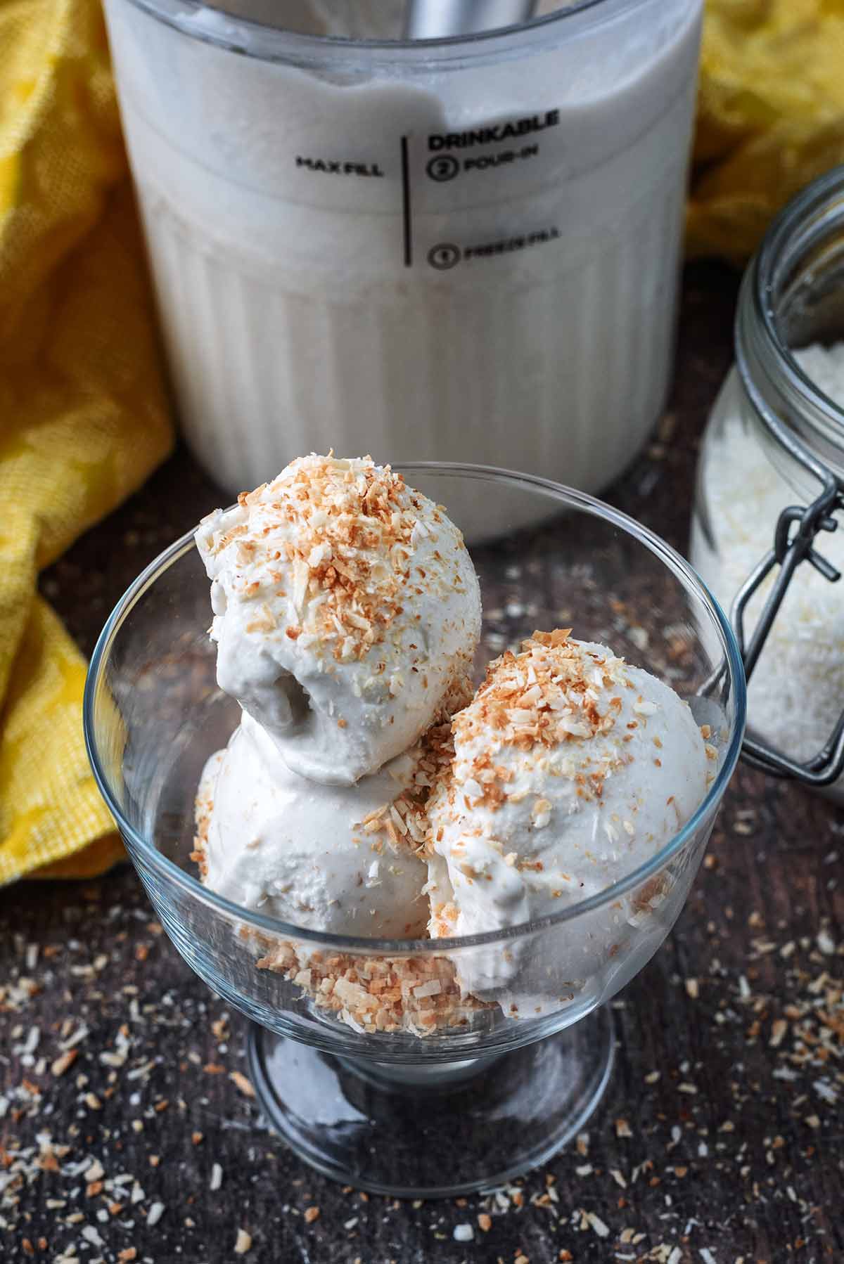 A serving glass full of white ice cream in front of a tub of more ice cream.