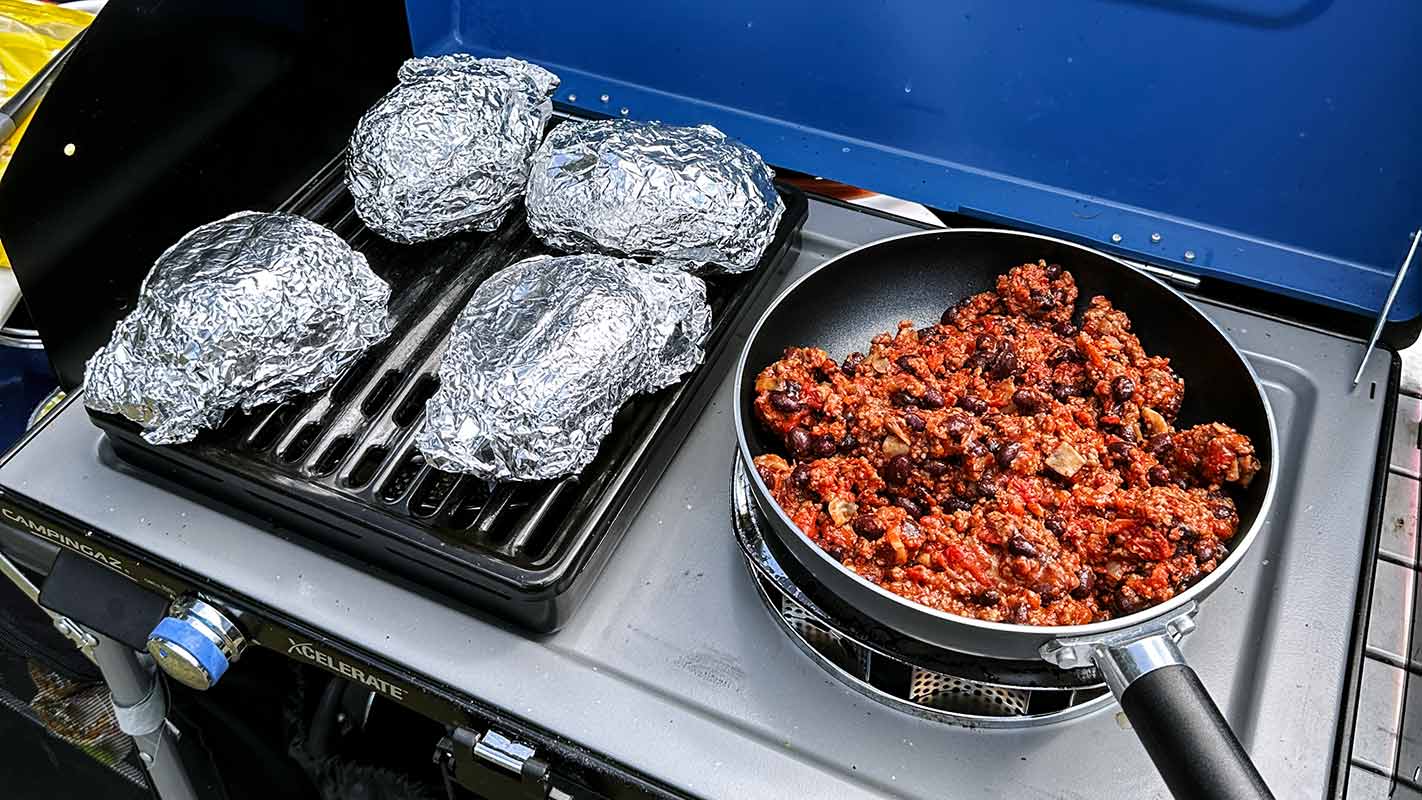 A pan of chilli cooking in a pan next to foil wrapped potatoes.