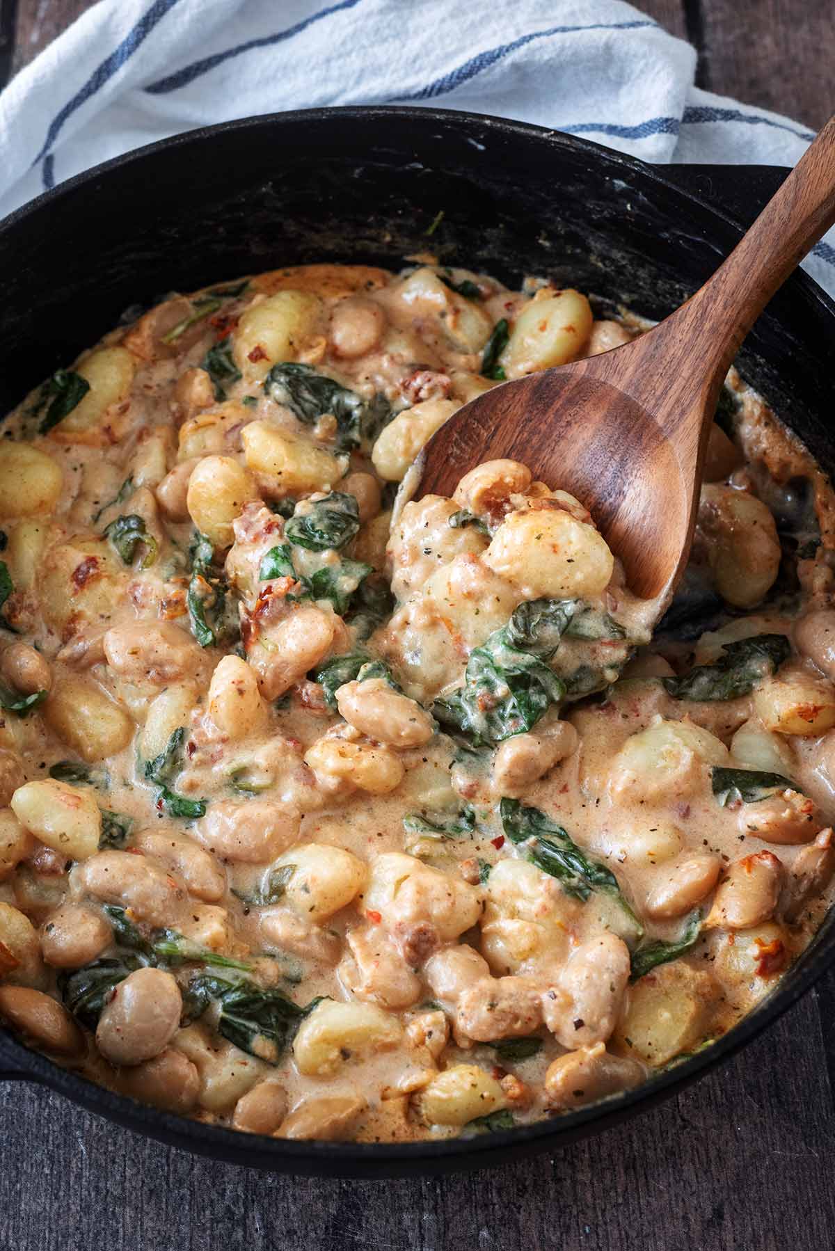 A wooden spoon lifting some gnocchi and beans out of a creamy sauce.