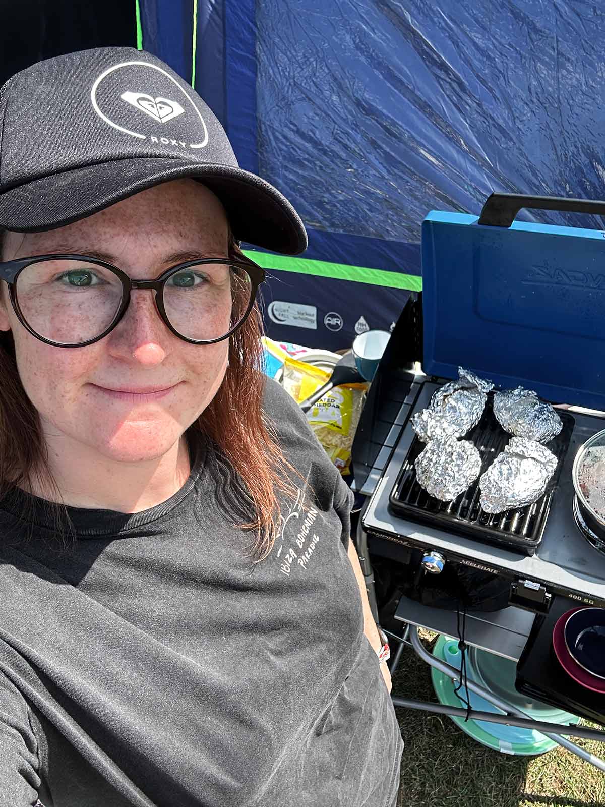A woman in hat and glasses stood in front of a camping stove.