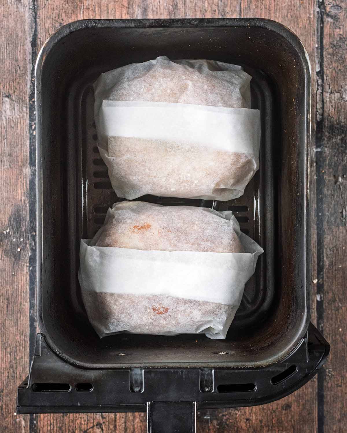 Two parchment paper wrapped bagels in an air fryer basket.
