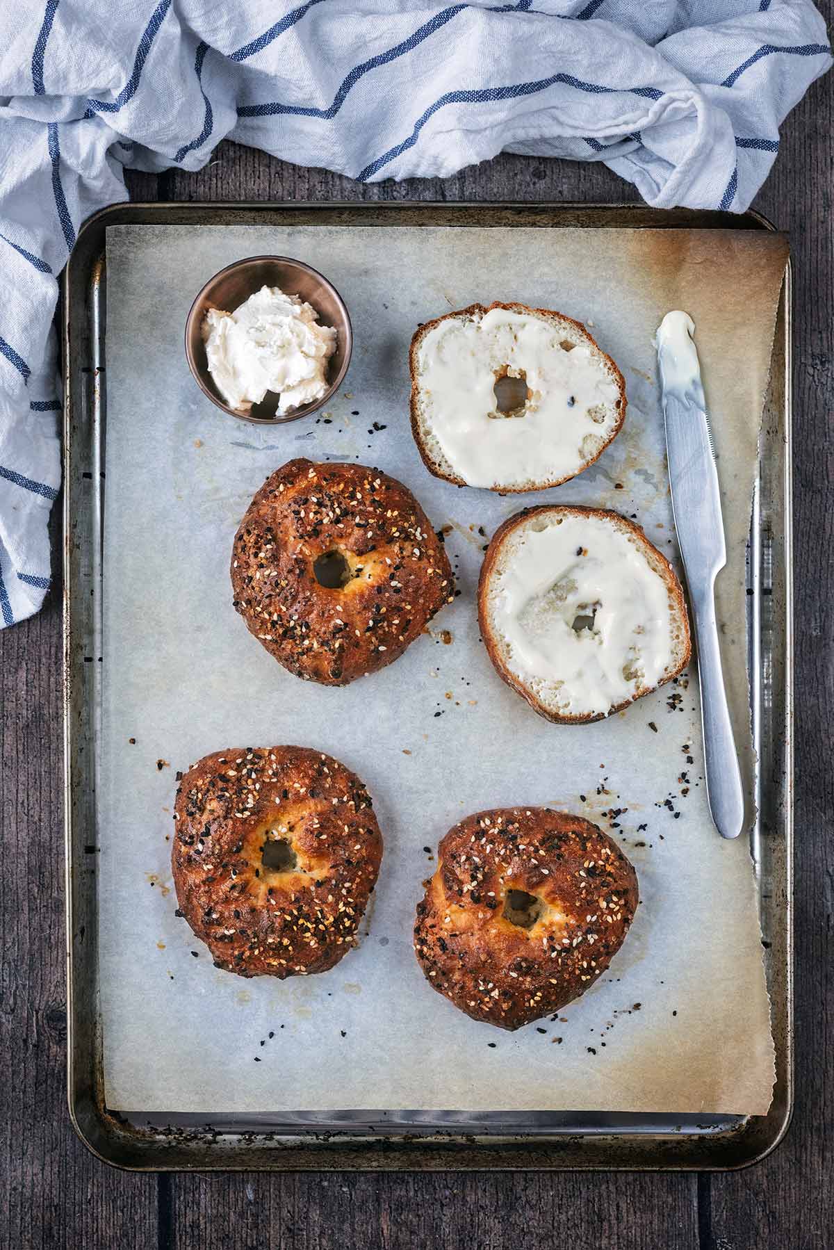 Four freshly baked bagels on a baking traym, one is sliced in half.