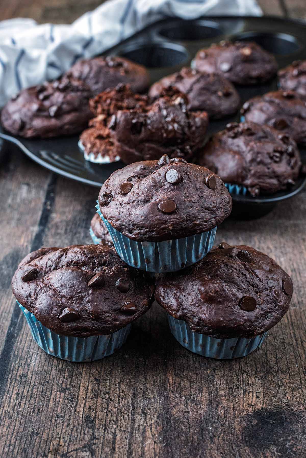 A stack of chocolate muffins in front of a baking tray full of more muffins.