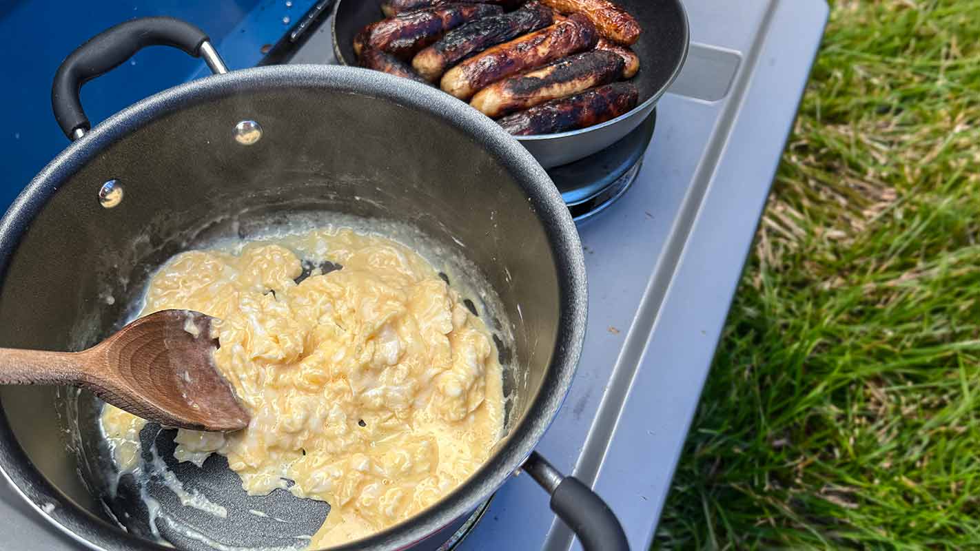 A pan of scrambled eggs next to a pan of sausages.
