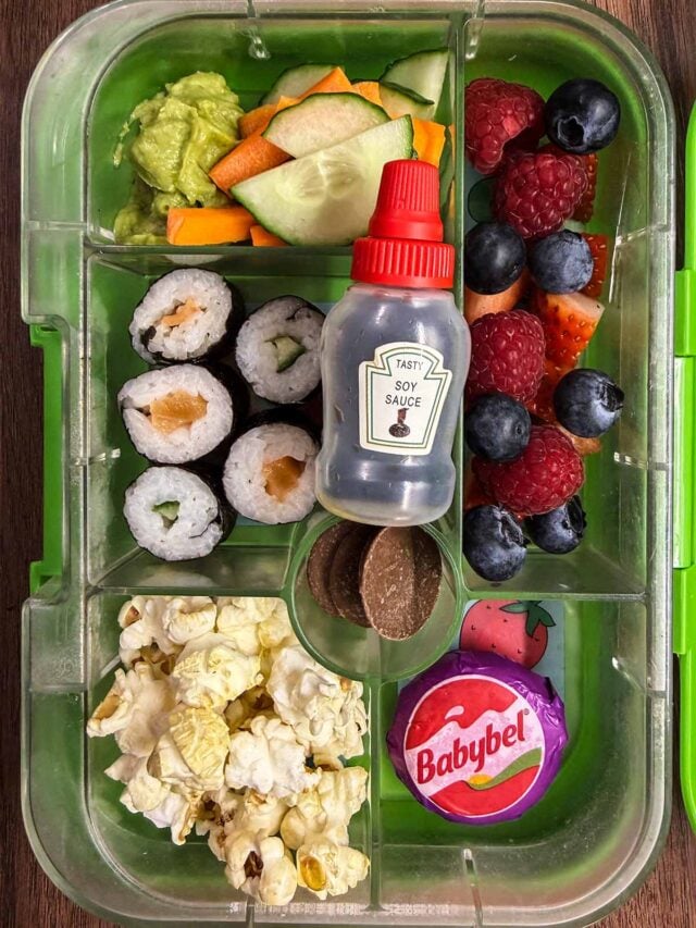 A compartment lunch box containing sushi, salad, popcorn, chocolate buttons, berries, cheese and a small bottle of soy sauce.