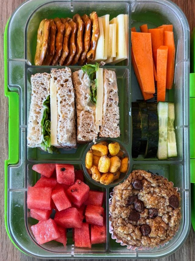 A compartment lunchbox containing a cheese salad sandwich, crackers, small cheese slices, carrot sticks, cucumber slices, corn, watermelon cubes and a chocolate chip muffin.