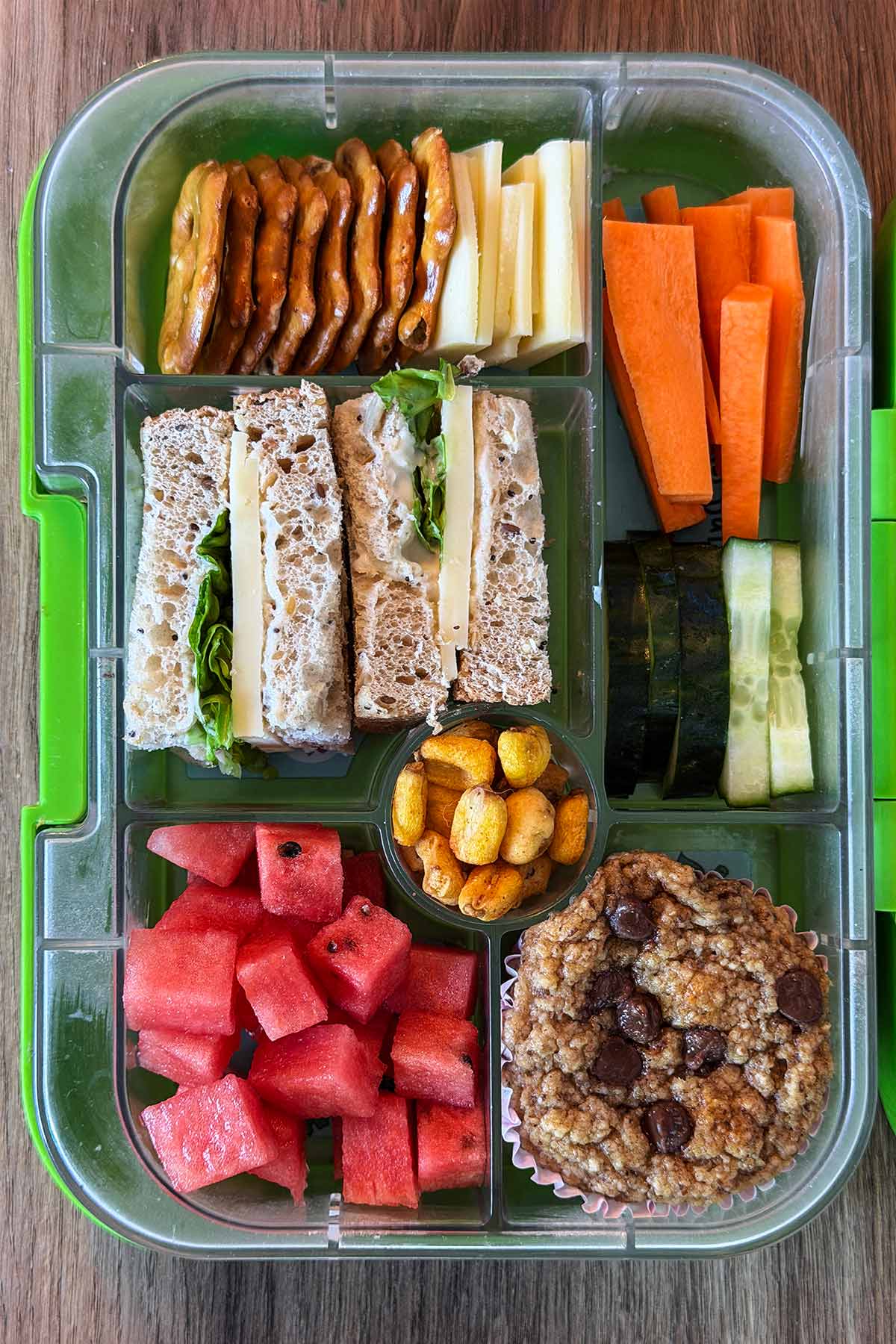A compartment lunchbox containing a cheese salad sandwich, crackers, small cheese slices, carrot sticks, cucumber slices, corn, watermelon cubes and a chocolate chip muffin.