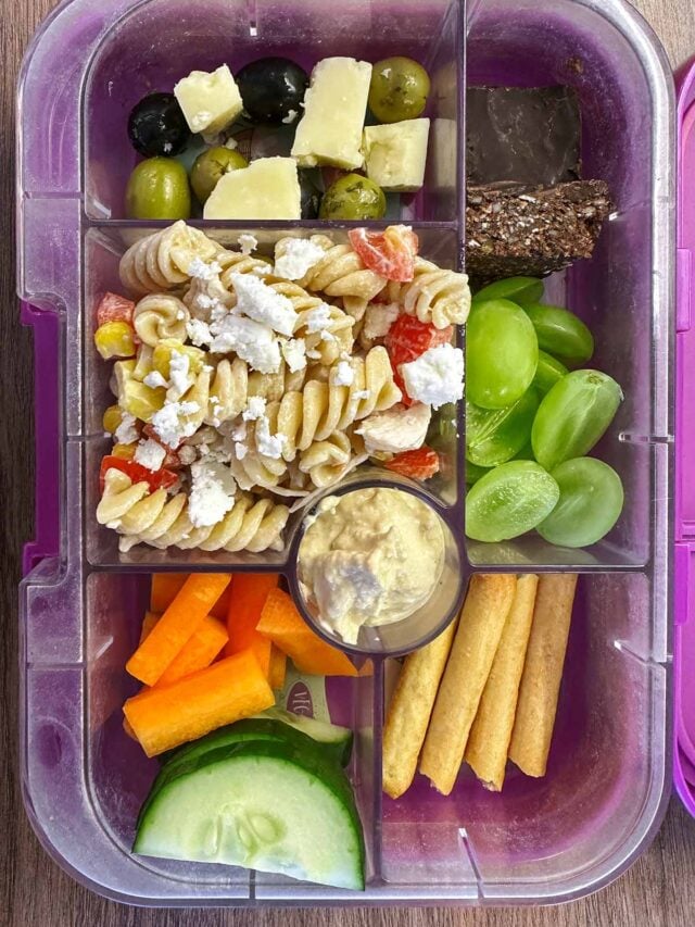 A compartment lunchbox containing pasta salad, cheese, olives, carrot sticks, cucumber slices, hummus, breadsticks, grapes and a chocolate seed bar.