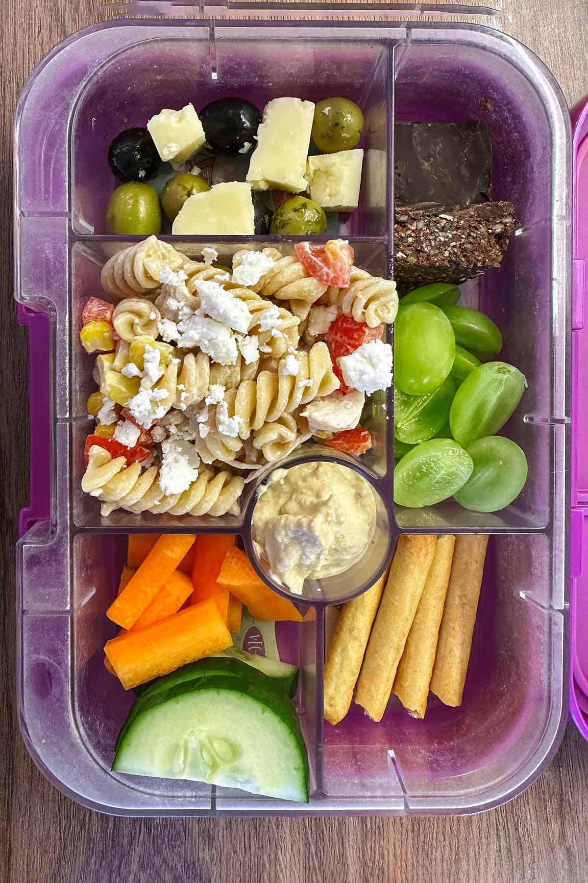 A compartment lunchbox containing pasta salad, cheese, olives, carrot sticks, cucumber slices, hummus, breadsticks, grapes and a chocolate seed bar.