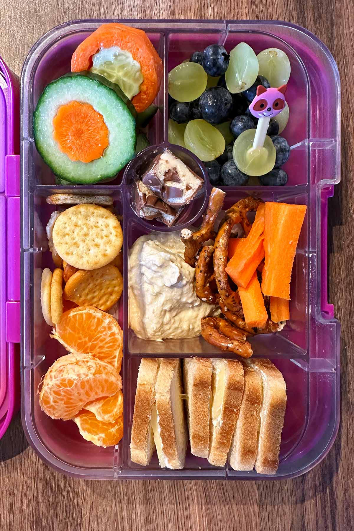 A compartment lunchbox containing sliced carrot and cucumbers, satsuma segments, grapes, blueberries, carrot sticks, hummus, mini pretzels and a cheese sandwich.