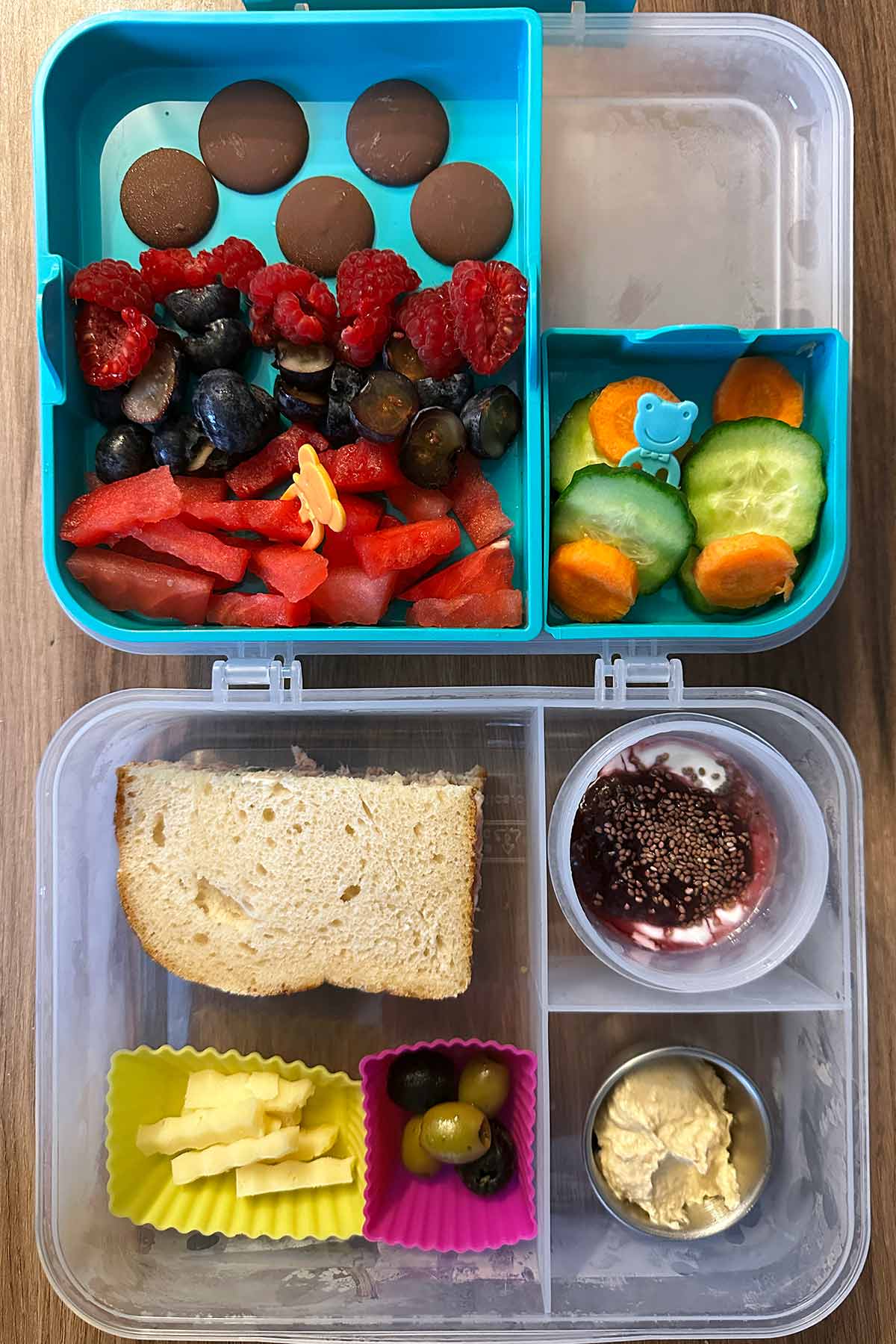 A compartment lunchbox containing a sandwich, berries, chocolate buttons, sliced carrot and cucumber, a pot of compote topped yogurt, a pot of hummus, a pot of cheese and a pot of olives.