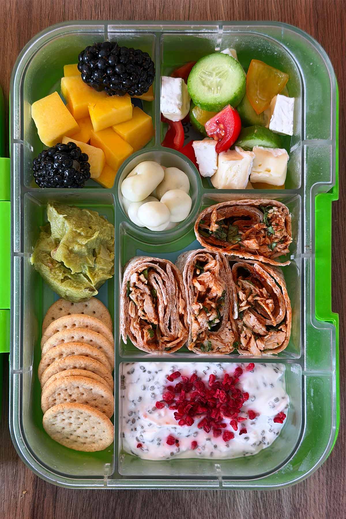 A compartment lunchbox containing chicken wraps, crackers, guacamole, fruit, vegetables and chia pudding.