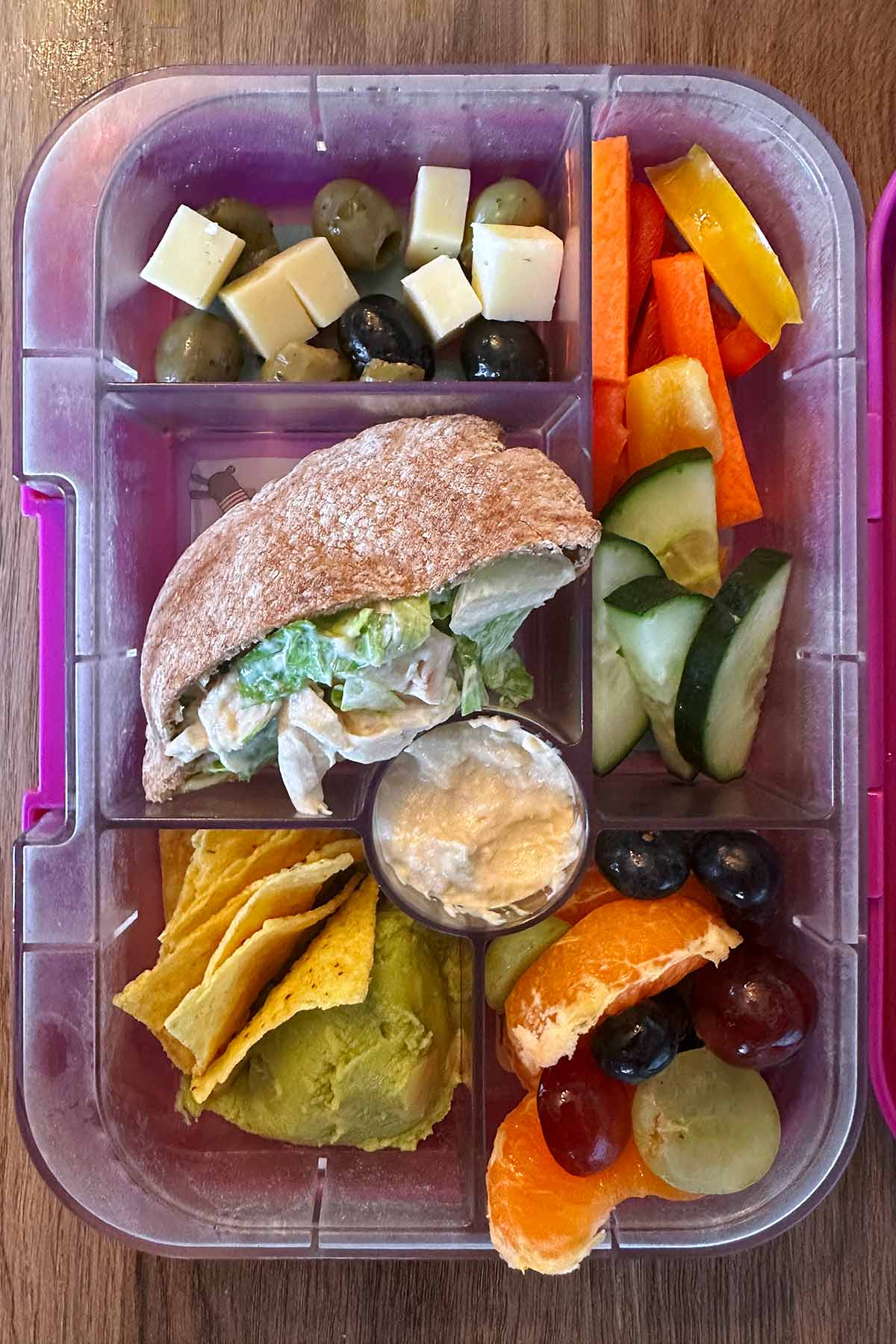 A compartment lunchbox containing a filled pita, guacamole and tortilla chips, sliced salad veg, cheese cubes, olives, grapes and satsuma segments.
