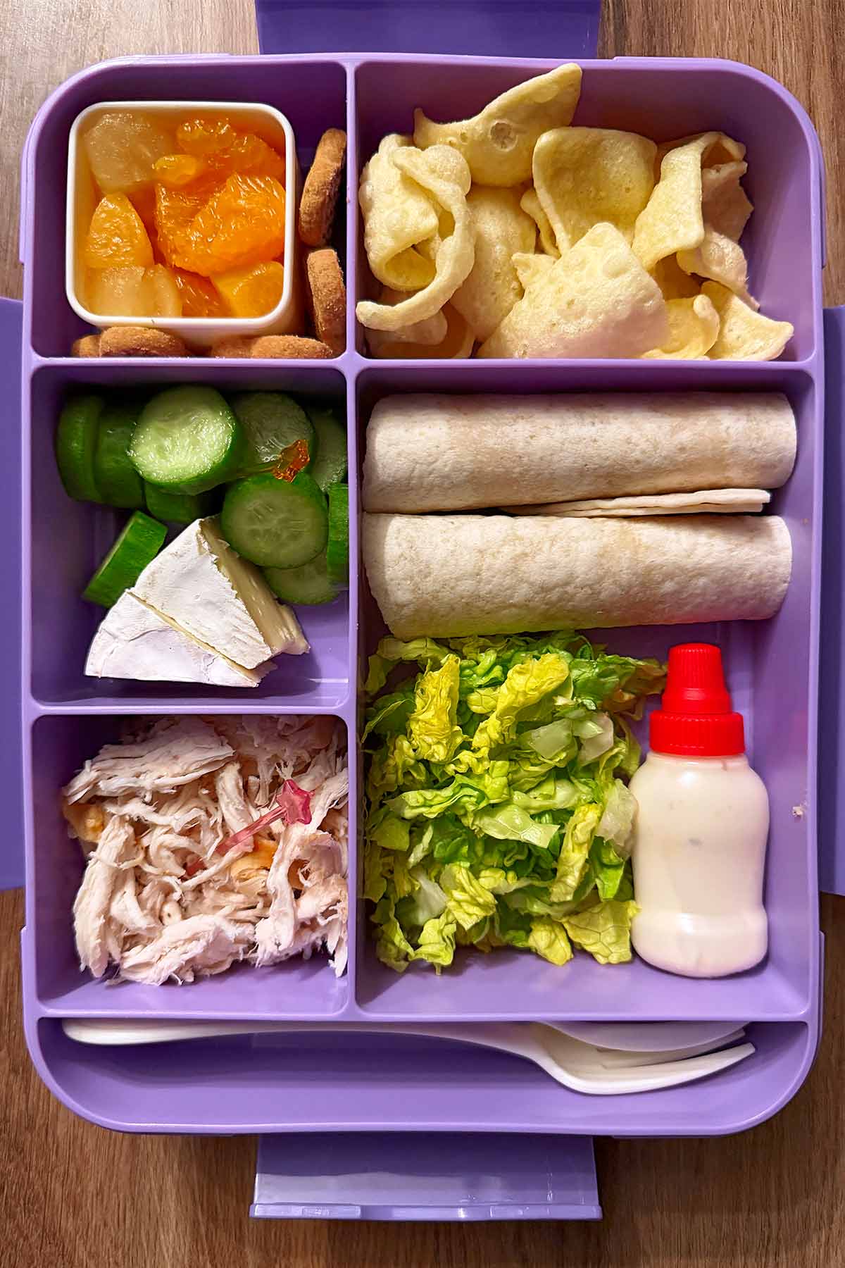 A compartment lunchbox containing a deconstructed chicken wrap, crisps, cheese, satsuma segments and mini cookies.