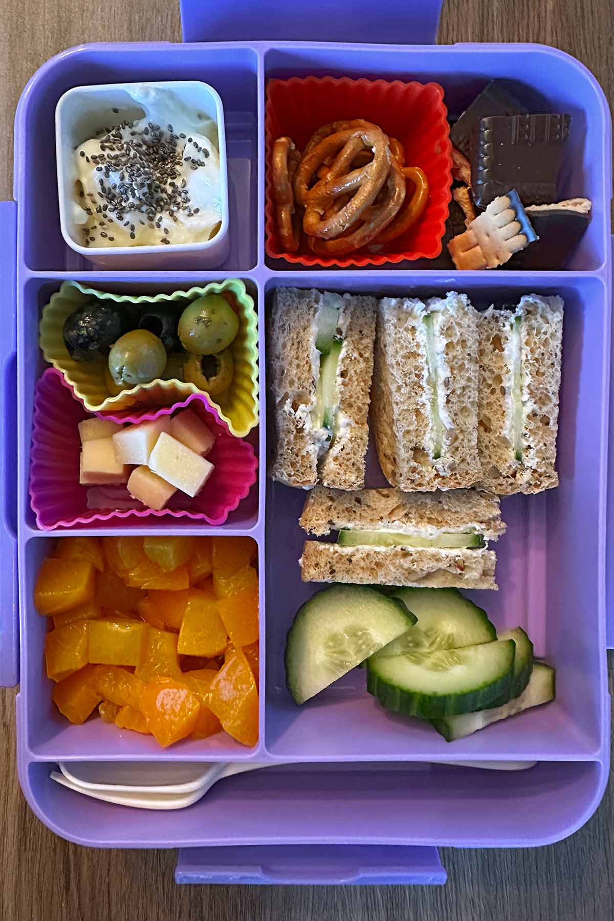 A compartment lunchbox containing a cucumber sandwich, a pot of yogurt, chocolate biscuit, cubes of peach, cucumber slices and silicone cups of cheese, olives and pretzels.