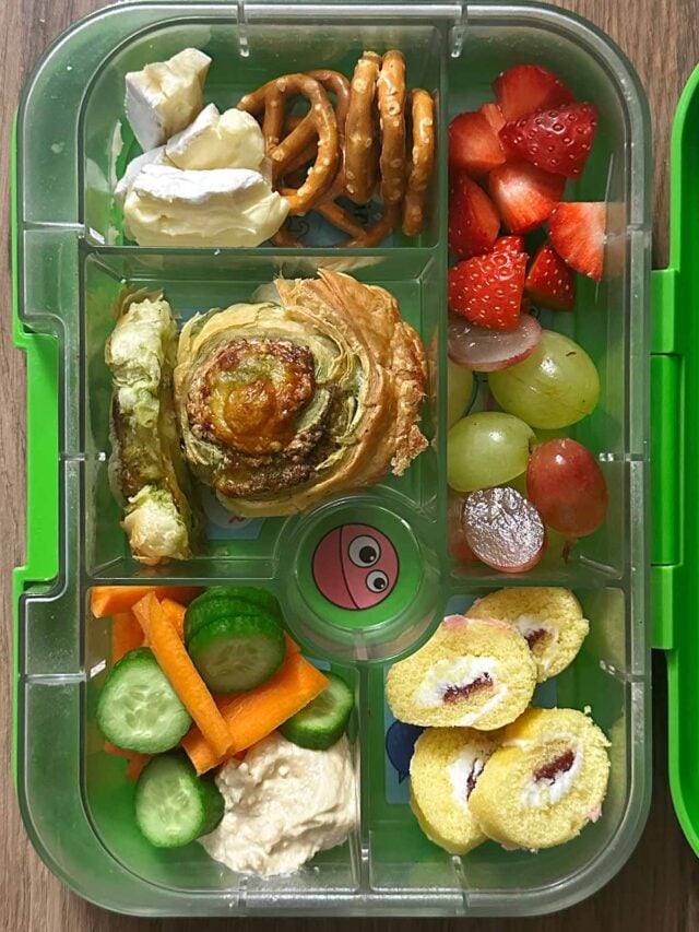 A compartment lunchbox containing a pinwheel, carrot, cucumber, hummus, berries, cheese and mini Swiss roll.
