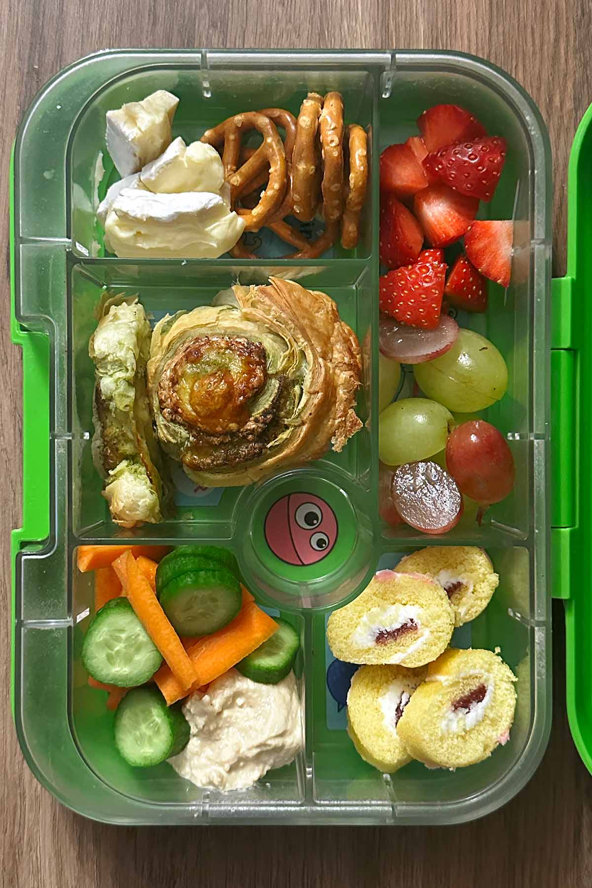 A compartment lunchbox containing a pinwheel, carrot, cucumber, hummus, berries, cheese and mini Swiss roll.