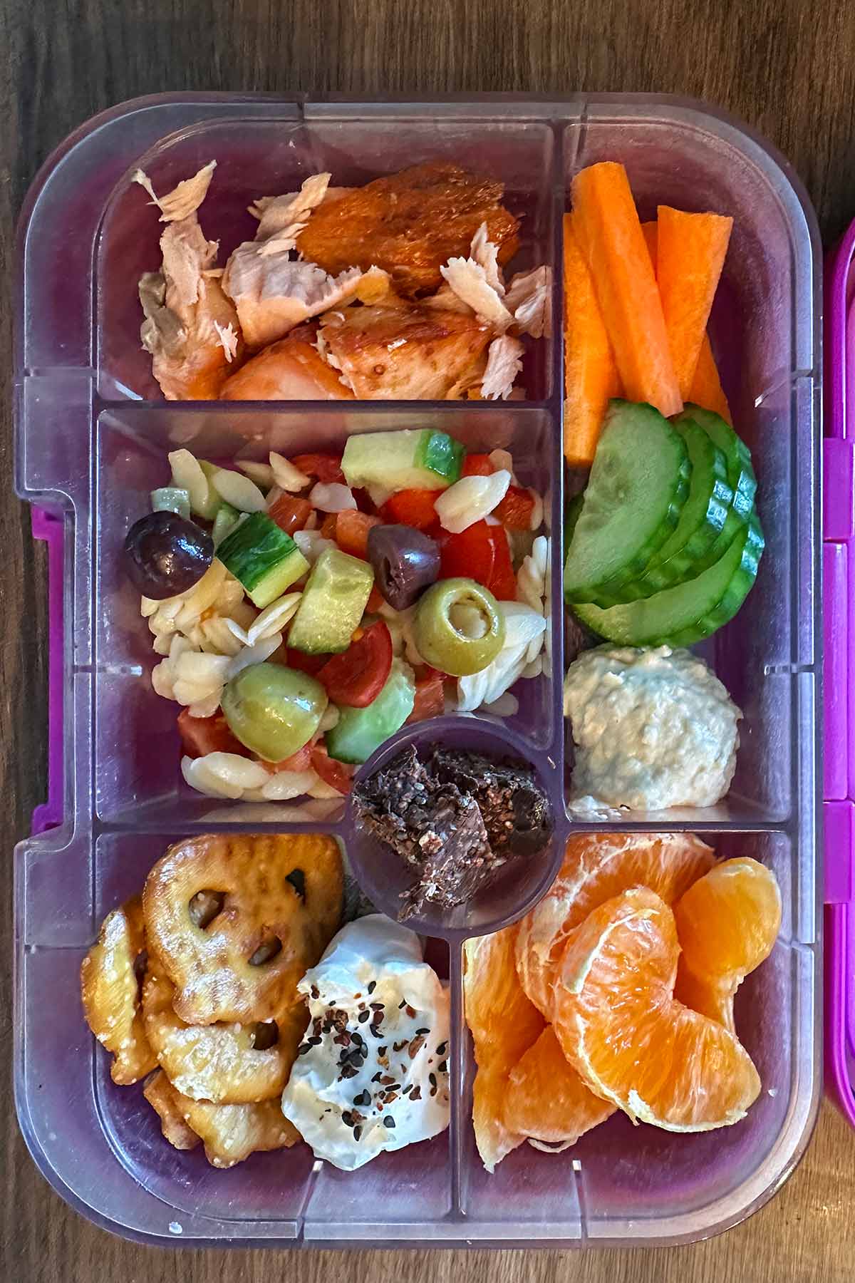 A compartment lunchbox containing flaked salmon, orzo salad, mini pretzels, hummus, crudités, cream cheese, orange segments and pieces of chocolate.