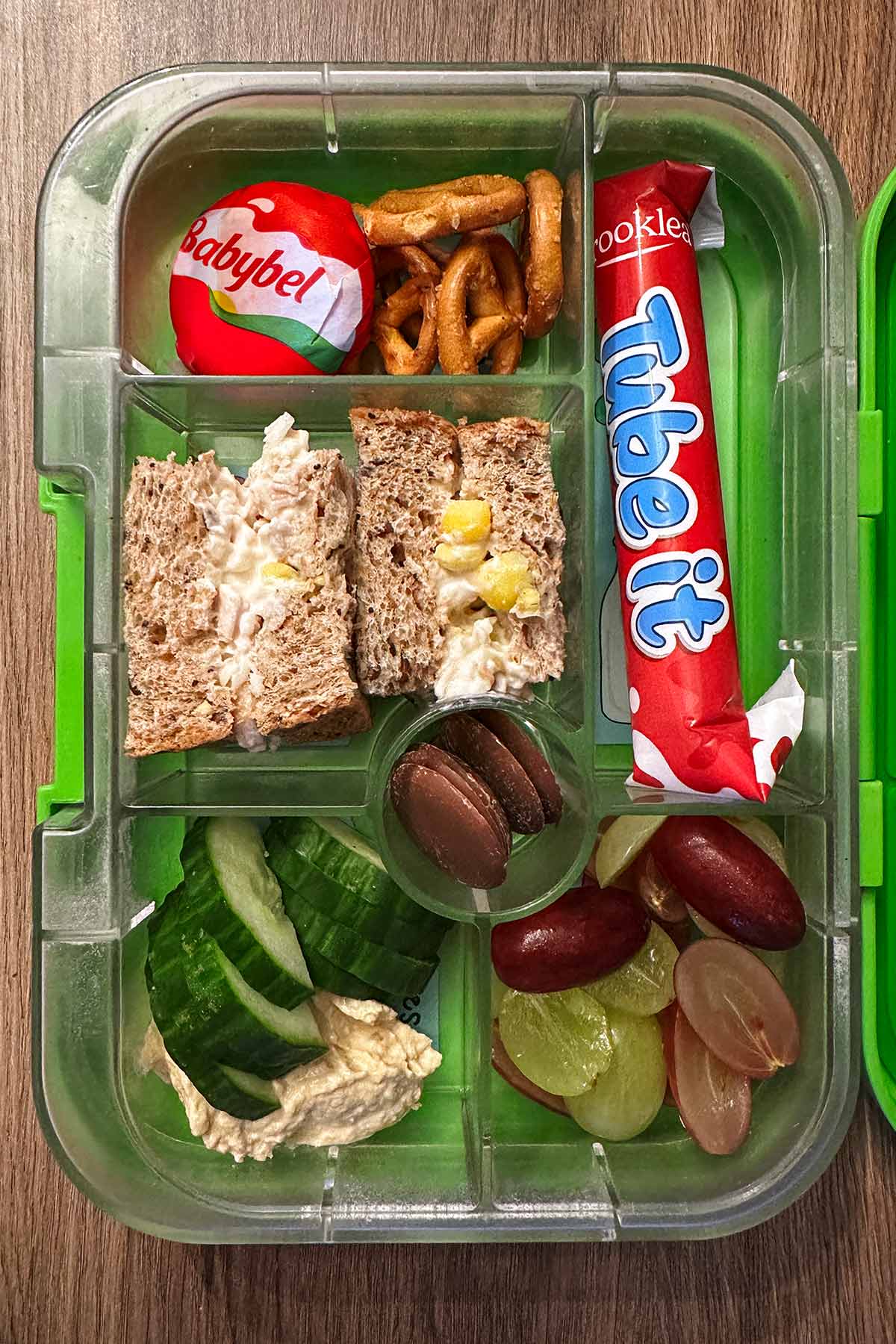 A compartment lunchbox containing a tuna mayo sandwich, cucumber, hummus, grapes, Babybel and a Frube yogurt.