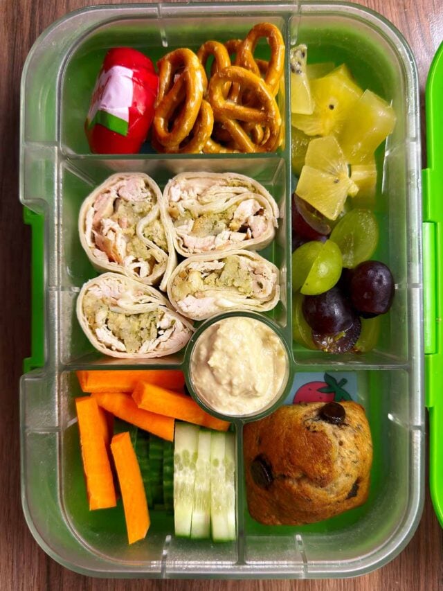 A compartment lunchbox containing wraps, carrots, cucumber, grapes, pretzels and a muffin.