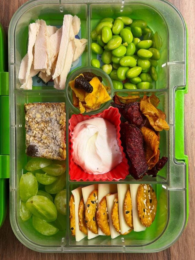 A compartment lunchbox containing shicken slices, crackers, cheese, edamame and grapes.