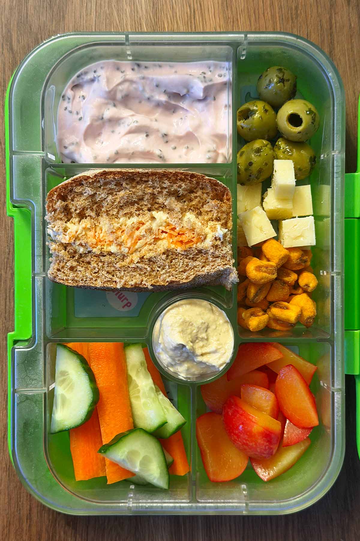 A compartment lunchbox containing a sandwich, olives, yogurt, carrot, cucumber, olives and corn.