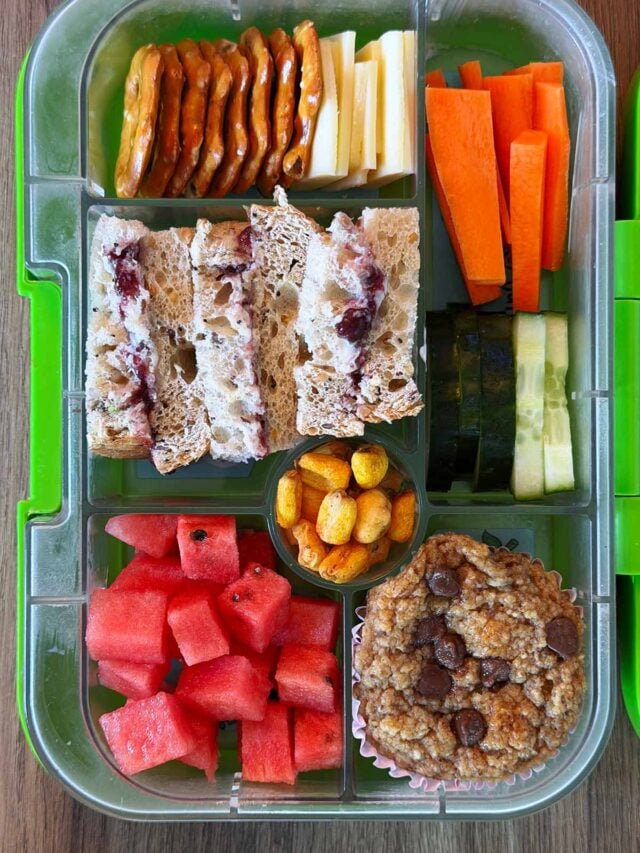A compartment lunchbox containing a sandwich, carrot sticks, crackers, cheese, watermelon and a muffin.