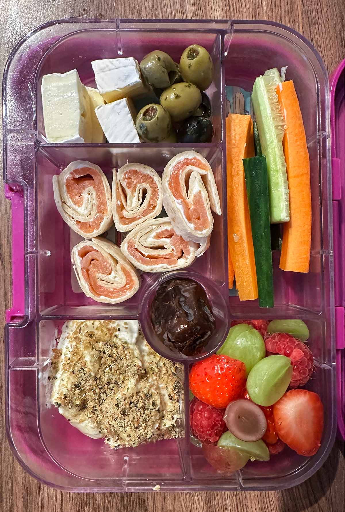 A compartment lunchbox containing salmon pinwheels, yogurt, brie, olives, crudités, berries and chocolate pudding.