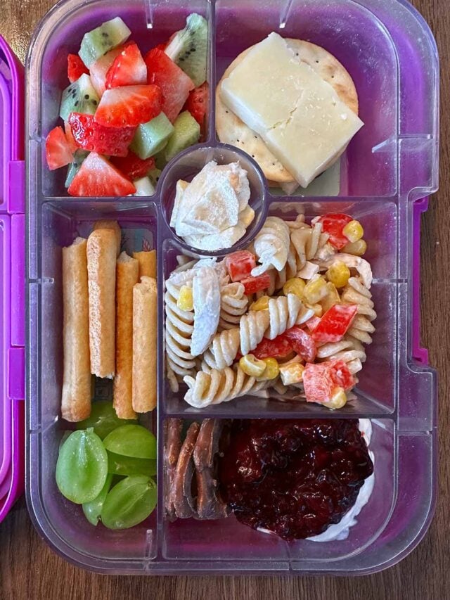 A compartment lunchbox containing pasta salad, cheese, crackers and fruit.