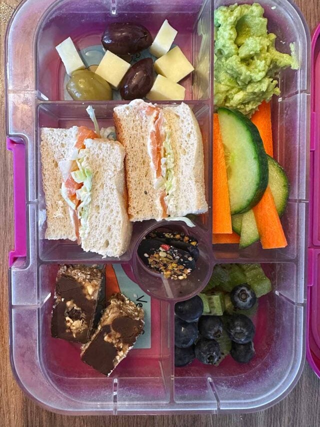 A compartment lunchbox containing a sandwich, guacamole, carrot, cucumber, fruit and flapjack.