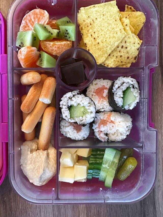 A compartment lunchbox containing sushi, tortilla chips, hummus, cucumber fruit and chocolate.