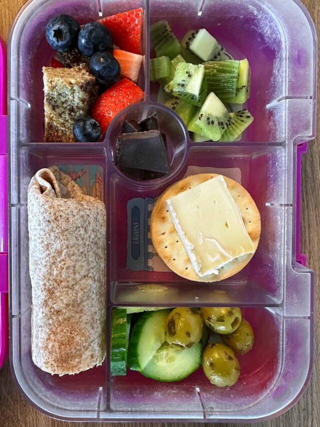 A compartment lunchbox containing a wrap, fruit, cheese and crackers, berries and a flapjack.