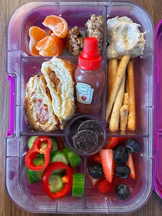 A compartment lunchbox containing sausage rolls, breadsticks, hummus and fruit.