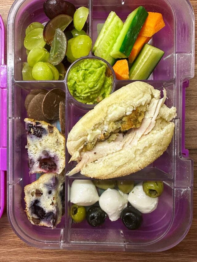 A compartment lunchbox containing a chicken roll, fruit, veg, cheese, chocolate and a muffin.