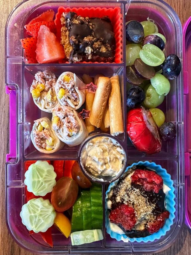 A compartment lunchbox containing tuna wrap, fruit, salad vegetables, breadsticks, hummus, fruit yogurt and flapjack.