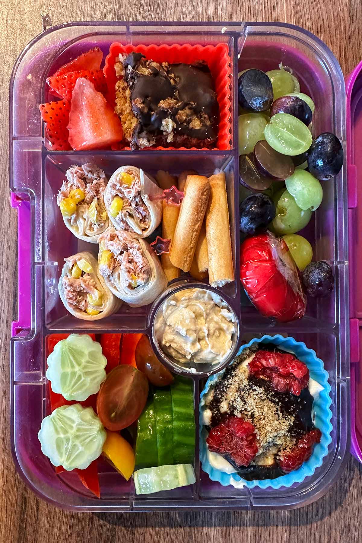 A compartment lunchbox containing tuna wrap, fruit, salad vegetables, breadsticks, hummus, fruit yogurt and flapjack.