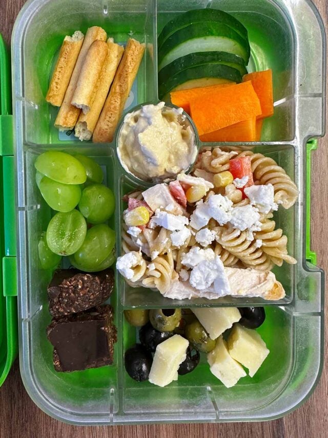 A compartment lunchbox containing pasta salad, cucumber, carrot, breadsticks, cheese and seed bars.