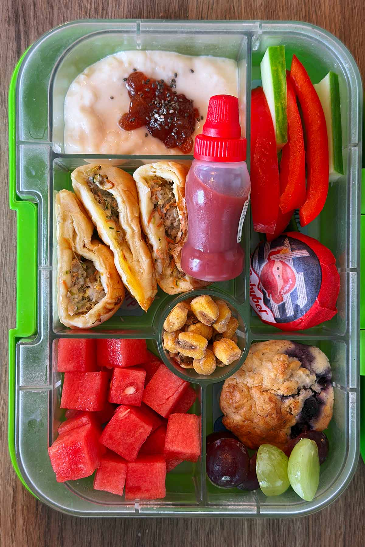 A compartment lunchbox containing sausage rolls, ketchup, cucumber, peppers, rice pudding, corn, melon and a muffin.