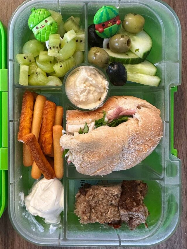 A compartment lunchbox containing a ham roll, kiwi, olives, cucumber, breadsticks, cream cheese and flapjack.