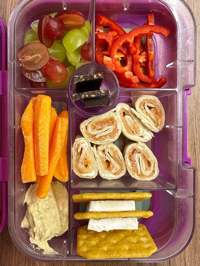 A compartment lunchbox containing salmon wrap, carrot sticks, cheese, crackers, grapes, hummus and chocolate buttons.