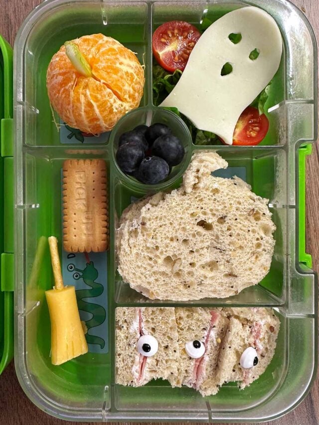 A compartment lunchbox containing a pumpkin shaped sandwich, ghost shaped cheese, satsuma, berries, tomatoes and a biscuit.