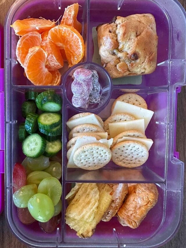 A compartment lunchbox containing cheese and crackers, cocktail sausages, crisps, fruit, veg and a muffin.